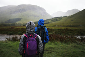 A couple hikes in full rain gear through an expansive, green terrain.